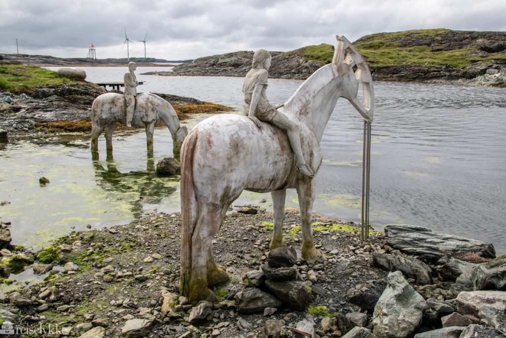 To skulpturer formet som hest og rytter i strandkanten, med fyrtårn og vindmøller i bakgrunnen